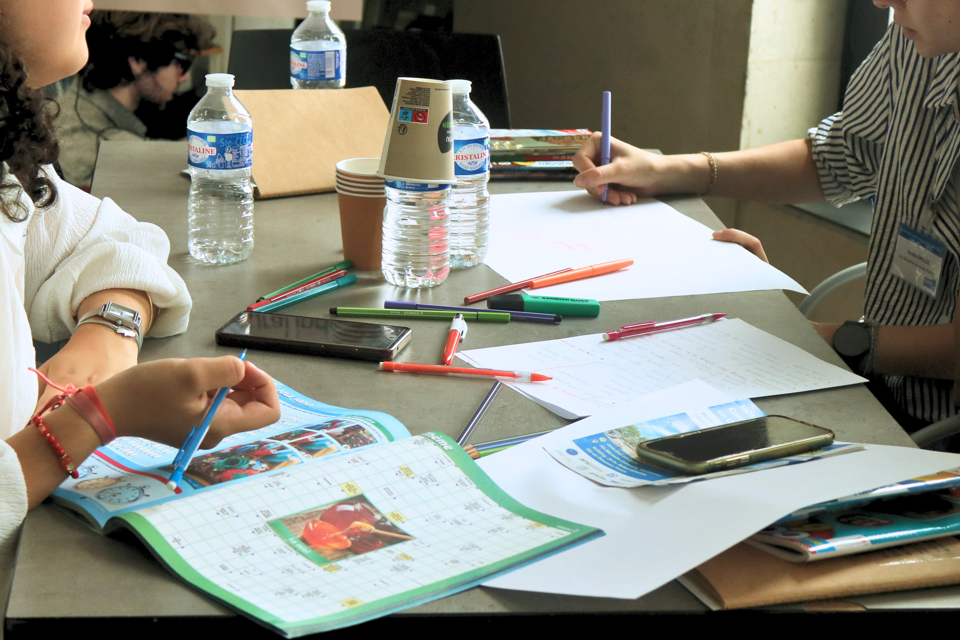 Une table sur laquelle sont posé des cahiers d'activité, des crayons et des bouteilles d'eau. 3 personnes y travaillent