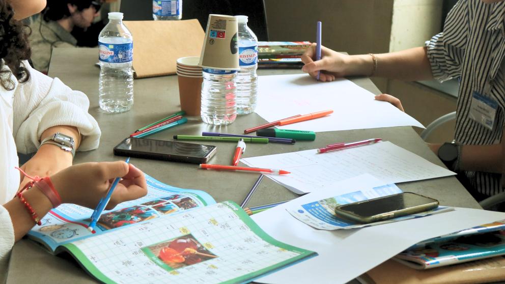 Une table sur laquelle sont posé des cahiers d'activité, des crayons et des bouteilles d'eau. 3 personnes y travaillent